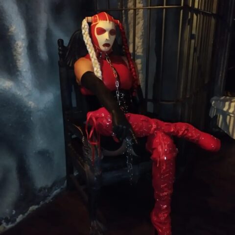 Eine Person mit einer dramatischen rot-weißen Maske, einem rot-schwarzen Kostüm und oberschenkelhohen, glänzenden roten Stiefeln sitzt auf einem dunklen Holzstuhl in einem schwach beleuchteten Raum mit blau-grauen, strukturierten Wänden.